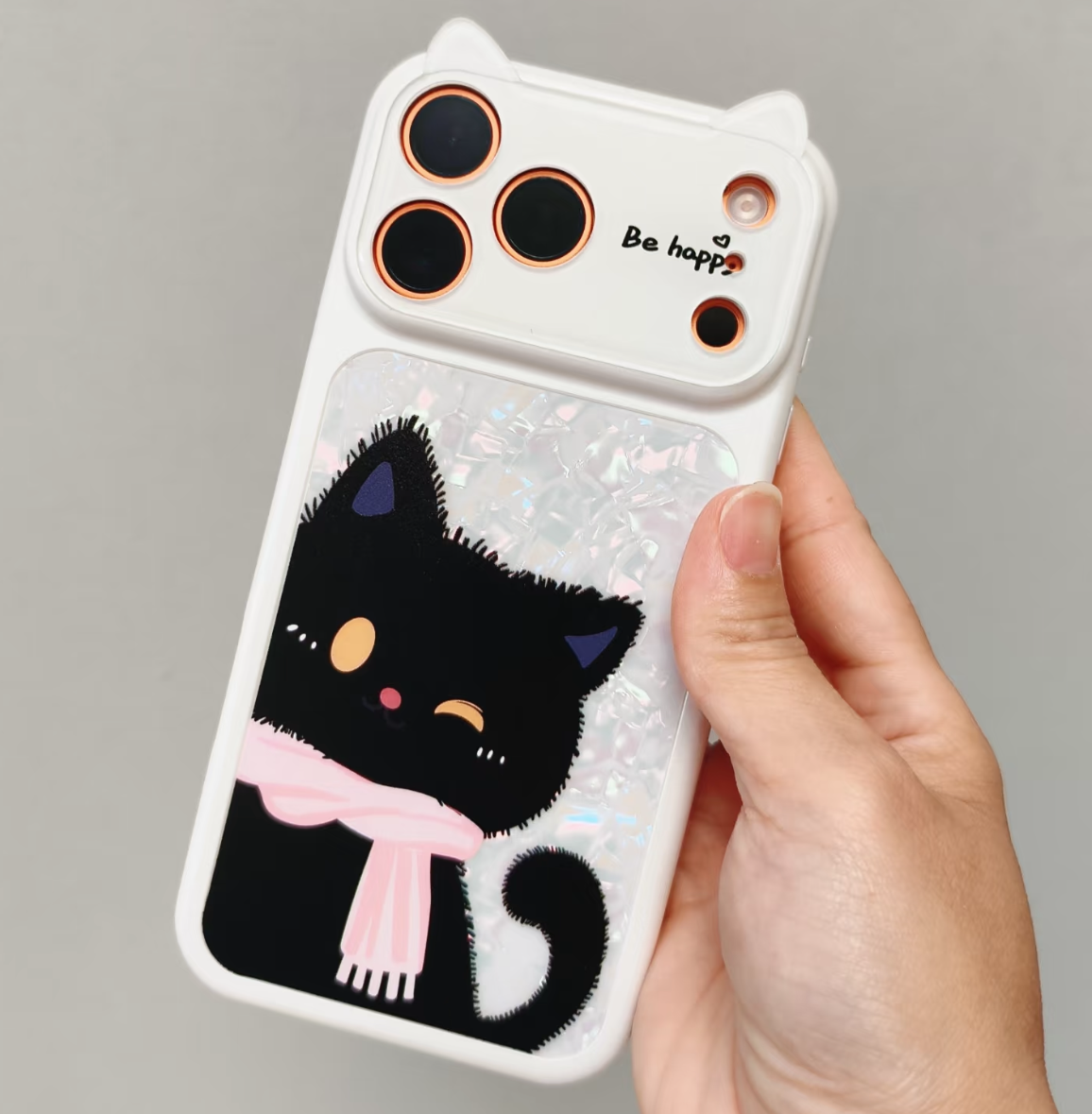 A hand holding a white phone case decorated with a black cartoon cat wearing a pink scarf, set against a plain gray background.